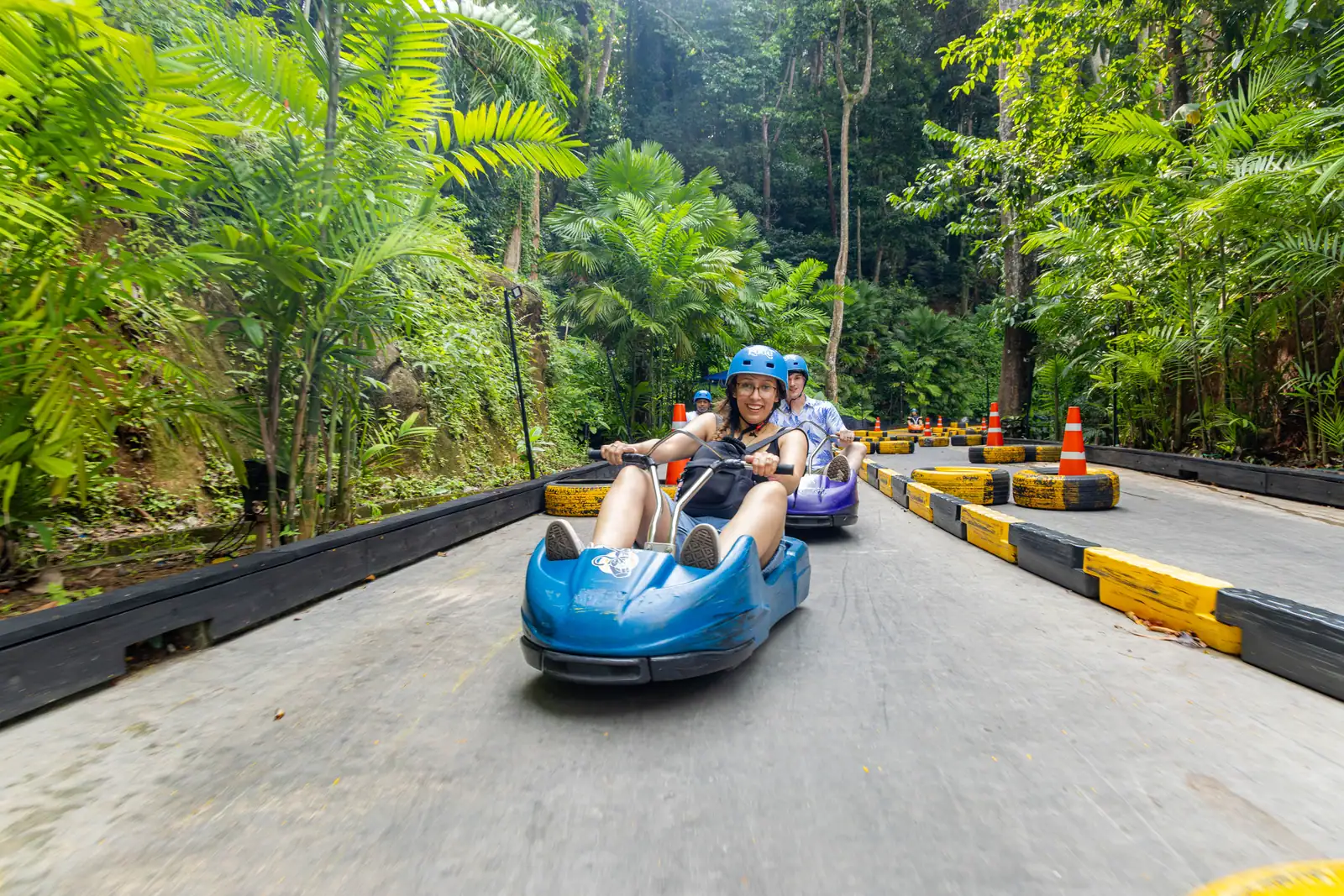 Helmeted riders in blue and purple luge carts on the jungle track.