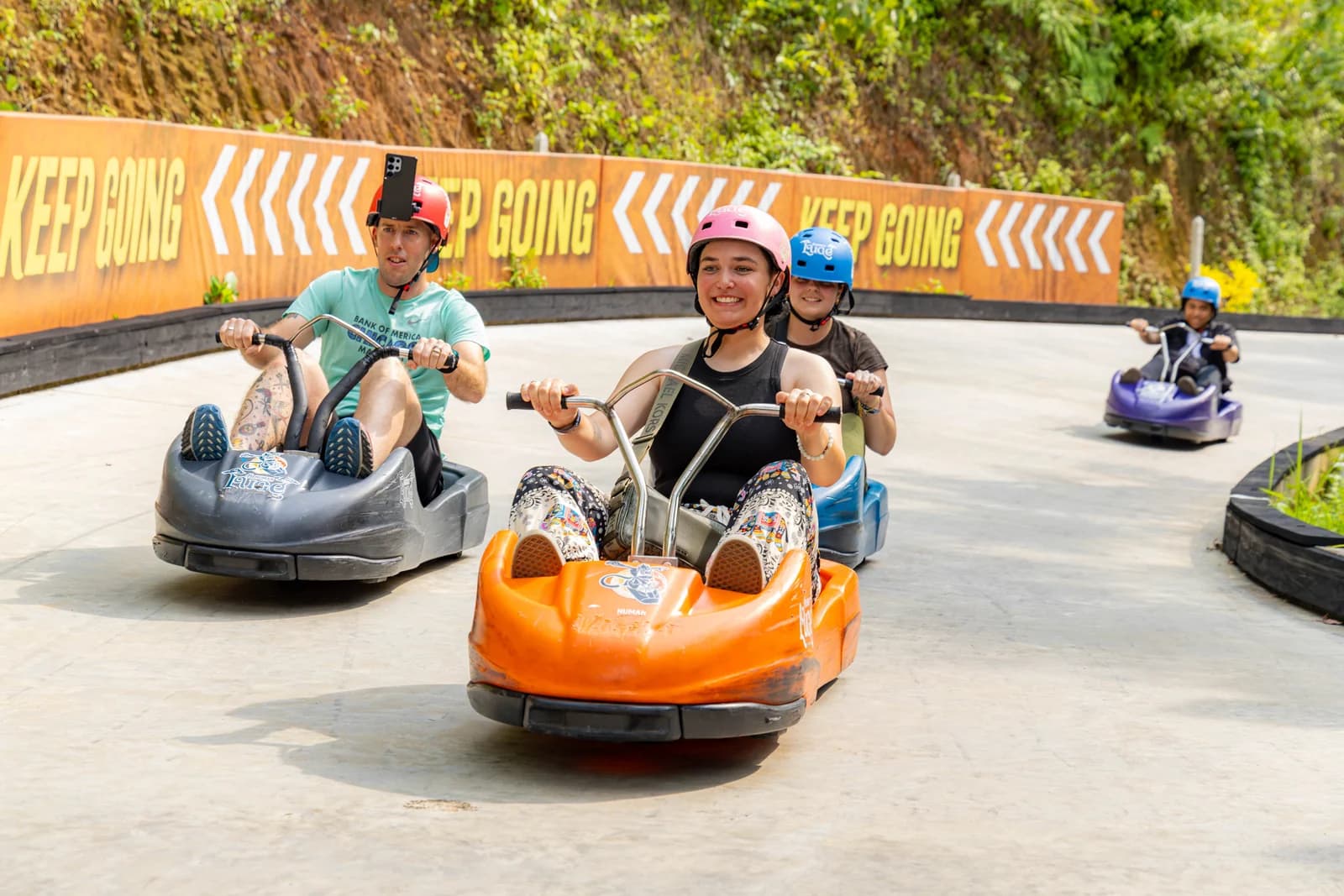 Helmeted riders in orange, black and blue luge carts rounding a bend.