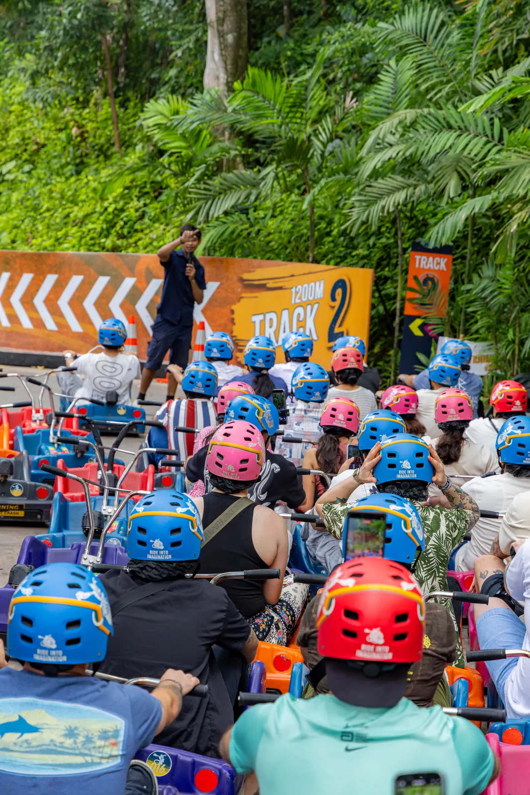 Helmeted riders seated in luge carts listening to a staff member near the Track 2 sign.
