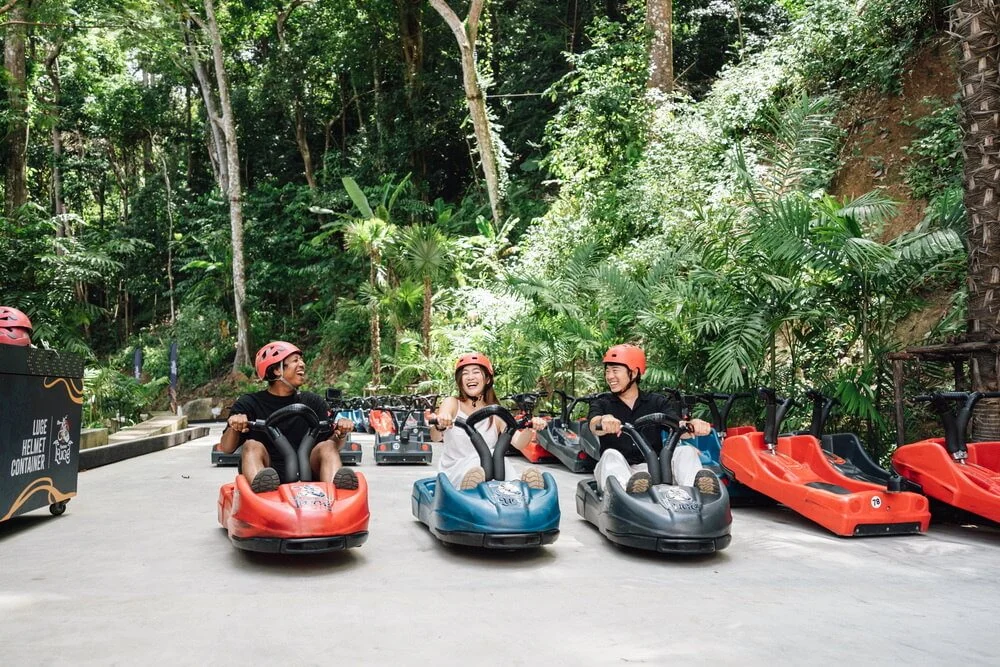 Three helmeted riders seated in separate luge carts beside the tropical track.